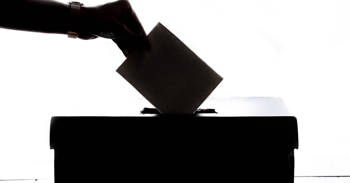 A person dropping a paper ballot into a wooden ballot box